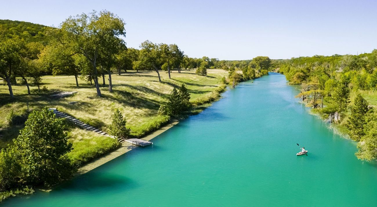 A kayaker paddling a clear stretch of the Blanco River, lined with green banks